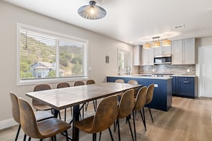 A modern kitchen with a central island, a dining area with a wooden table and chairs, and large windows with blinds. - Renovated 6BR GWS Escape Near Aspen - A/C (Glenwood Springs)