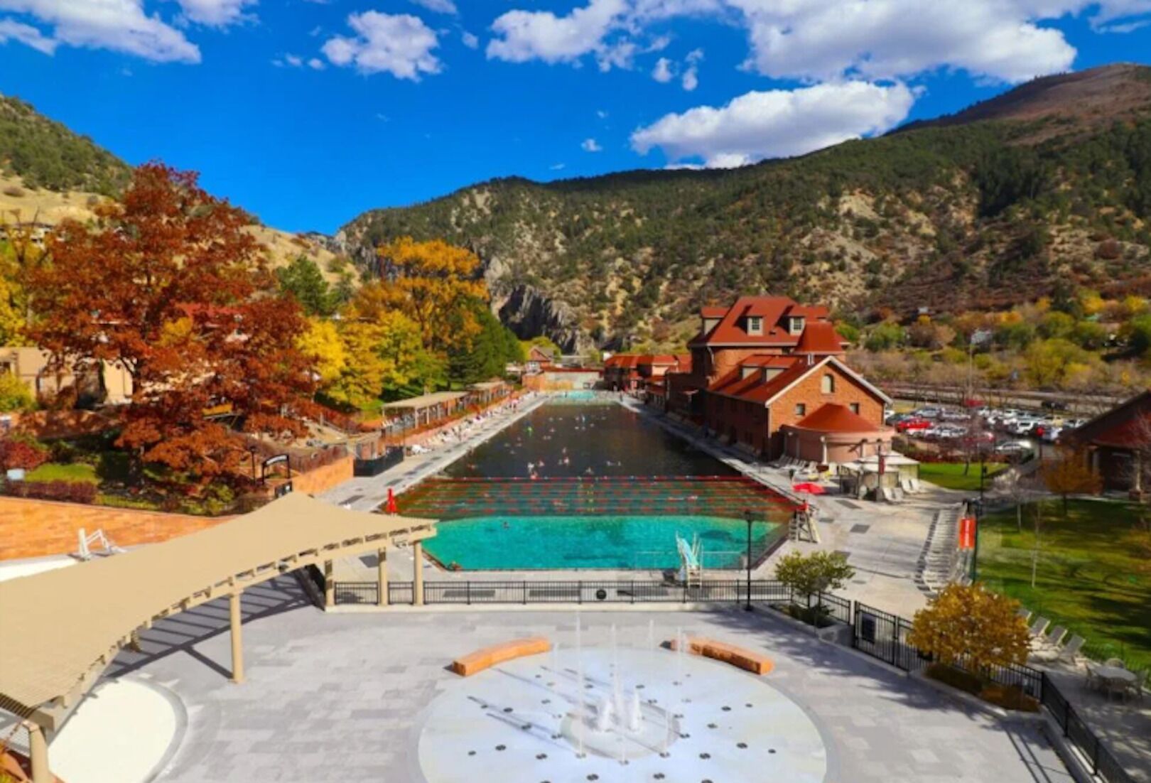 A swimming pool with a surrounding deck and a fountain, set against a backdrop of mountains and autumn foliage.