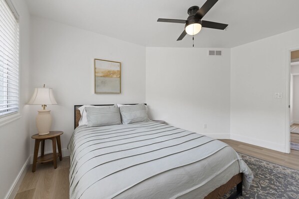 A bedroom with a bed, a ceiling fan, a lamp, and a painting on the wall.