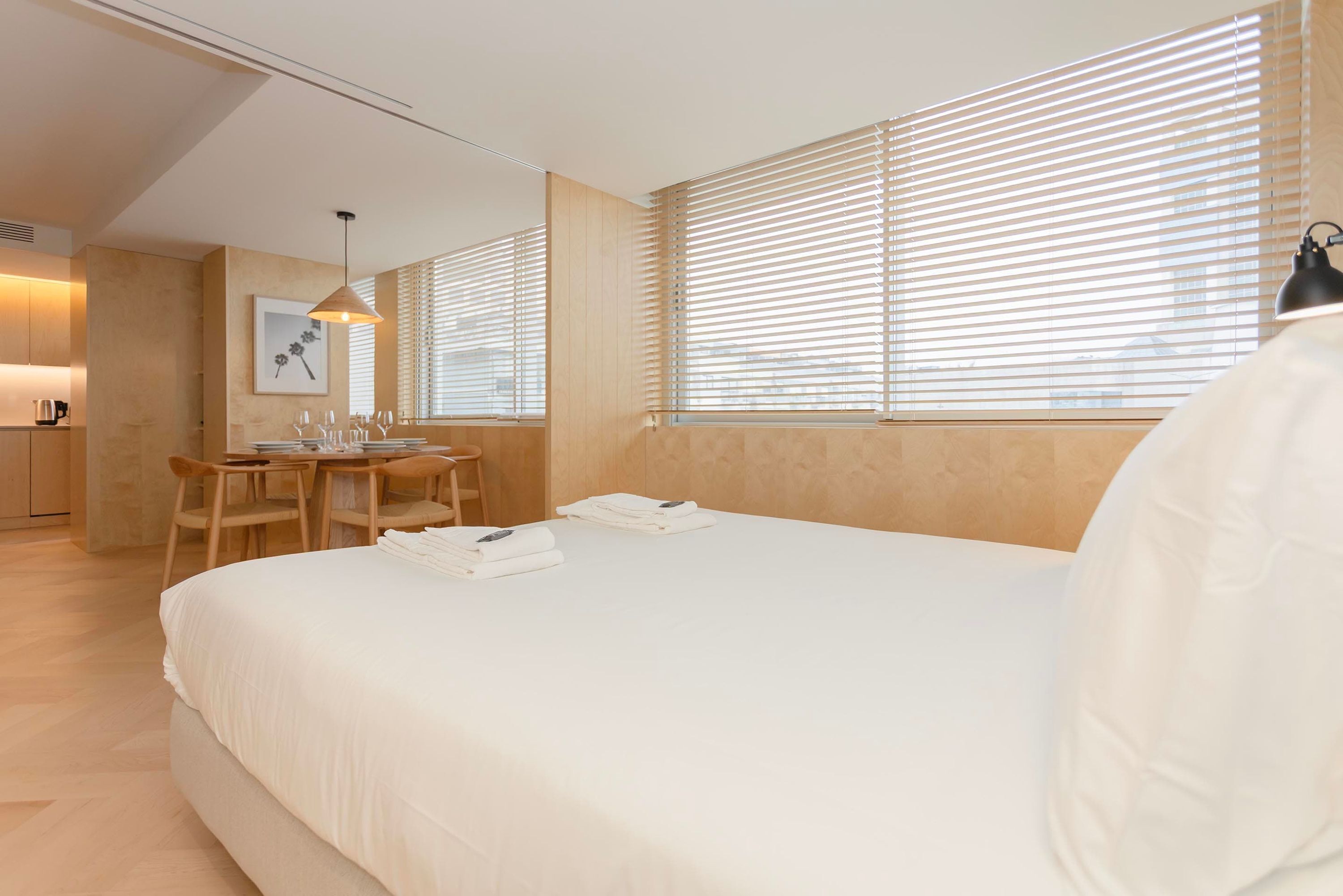 A hotel room with a large bed, a dining table with chairs, a kitchen area, and large windows with blinds.