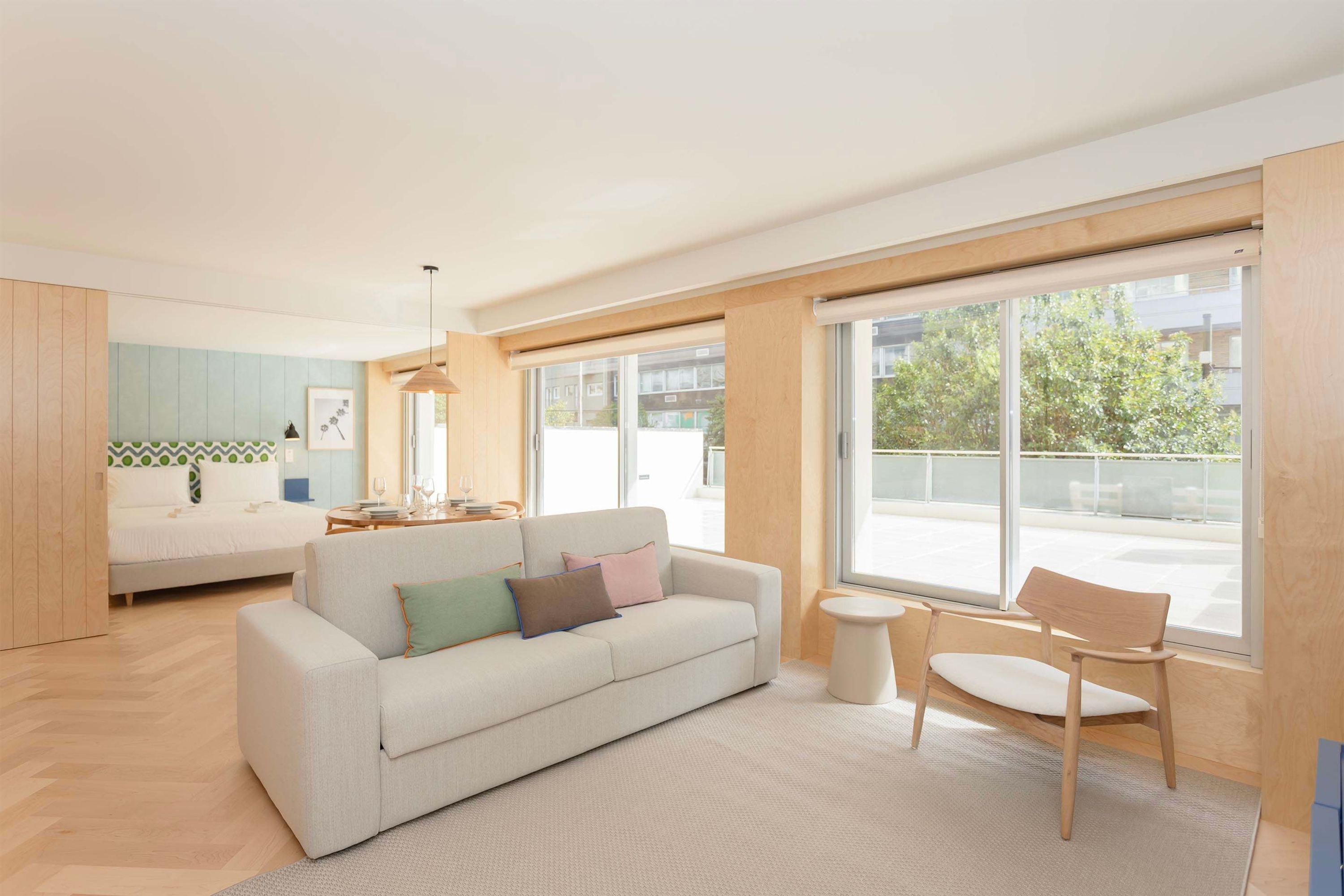 A modern living room with a sofa, a dining table, and a chair. There is a large window providing a view of the outside.