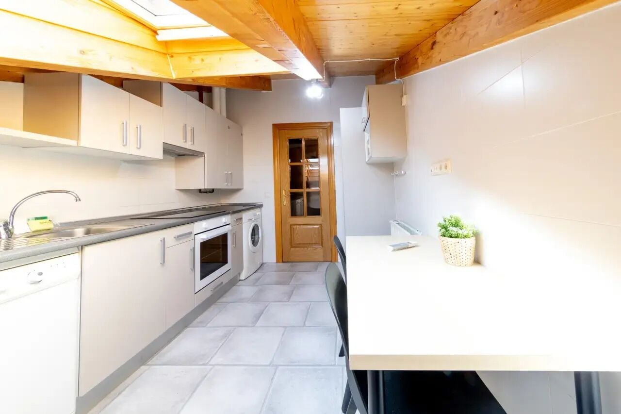 A compact kitchen with a wooden ceiling, a built-in oven, a washing machine, and a small dining area with a table and chairs.