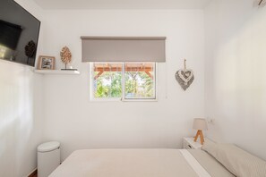 Ein Schlafzimmer mit einem Bett, einem Fenster mit Blick auf Grünflächen, einer Nachttischlampe und einem kleinen weißen Mülleimer.