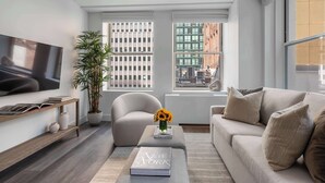 Un salon moderne doté d’un téléviseur à écran plat, d’un canapé, d’une table basse avec un livre, d’une plante en pot et de grandes fenêtres offrant une vue sur la ville.