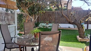 An outdoor patio with a glass table and metal chairs, surrounded by greenery and a decorative tree.