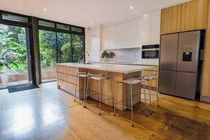 A modern kitchen with a large island, stainless steel appliances, and a view of a garden through the glass door. - South Coogee Sanctuary | Pool, Space & Sea Air (South Coogee)
