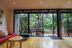 A modern interior with a red sofa, a small round table, and a bench. Large glass doors lead to a lush garden with a wooden table and chairs. - South Coogee Sanctuary | Pool, Space & Sea Air (South Coogee)