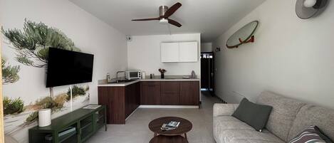 A modern living room with a flat-screen TV, a sofa, a coffee table, and a kitchen area with a microwave.