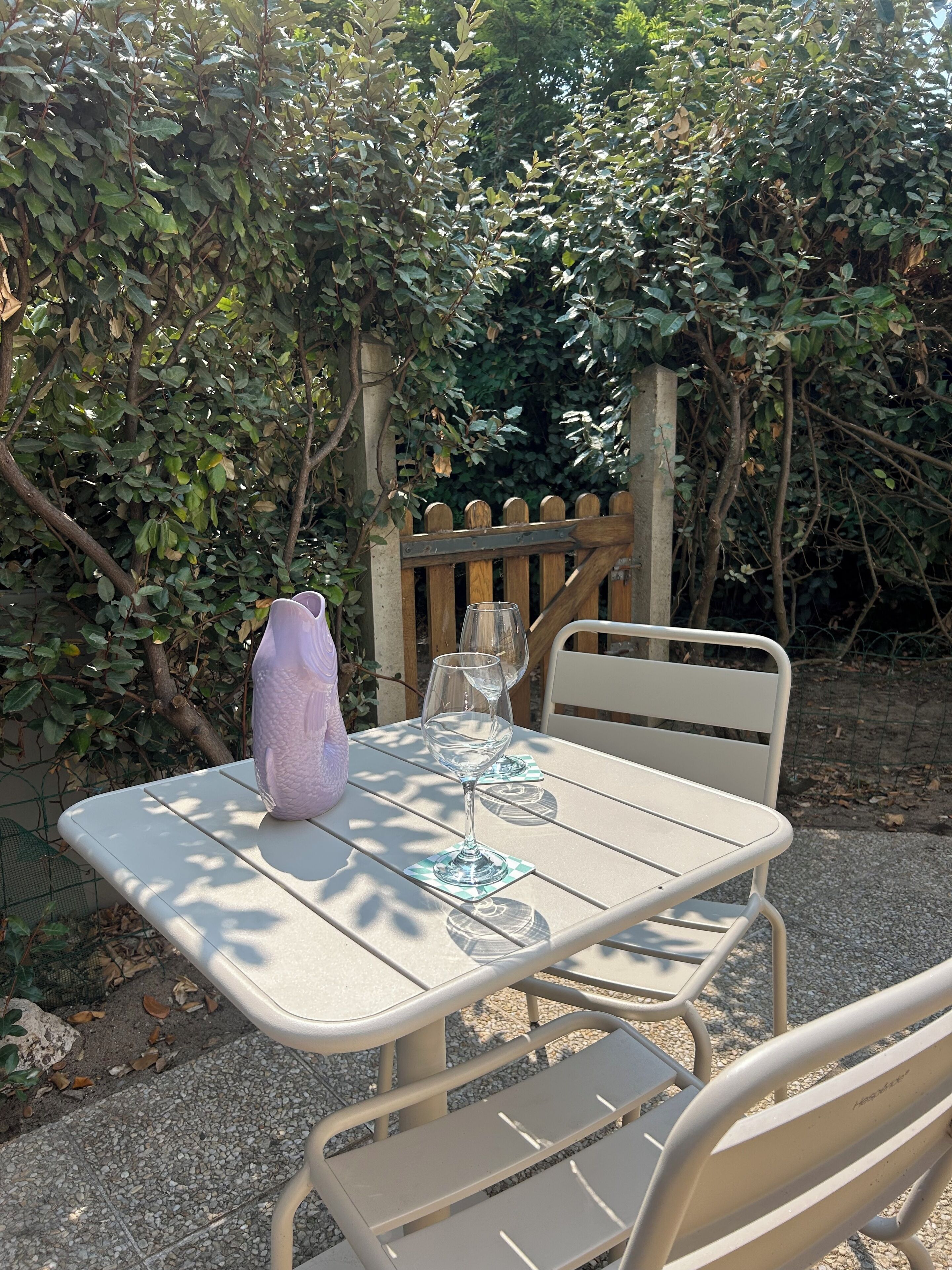 An outdoor table with a glass and a vase, set against a backdrop of greenery and a wooden fence.