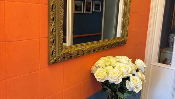 Espejo con un marco dorado ornamentado en una pared, un jarrón con flores blancas y una pared azul y naranja.