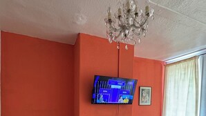 Una habitación con televisor montado en la pared, chimenea debajo, una tetera blanca sobre una mesa y una ventana con cortinas.