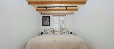 Un dormitorio con una cama, una ventana, un cuadro enmarcado en la pared y un techo con vigas de madera.