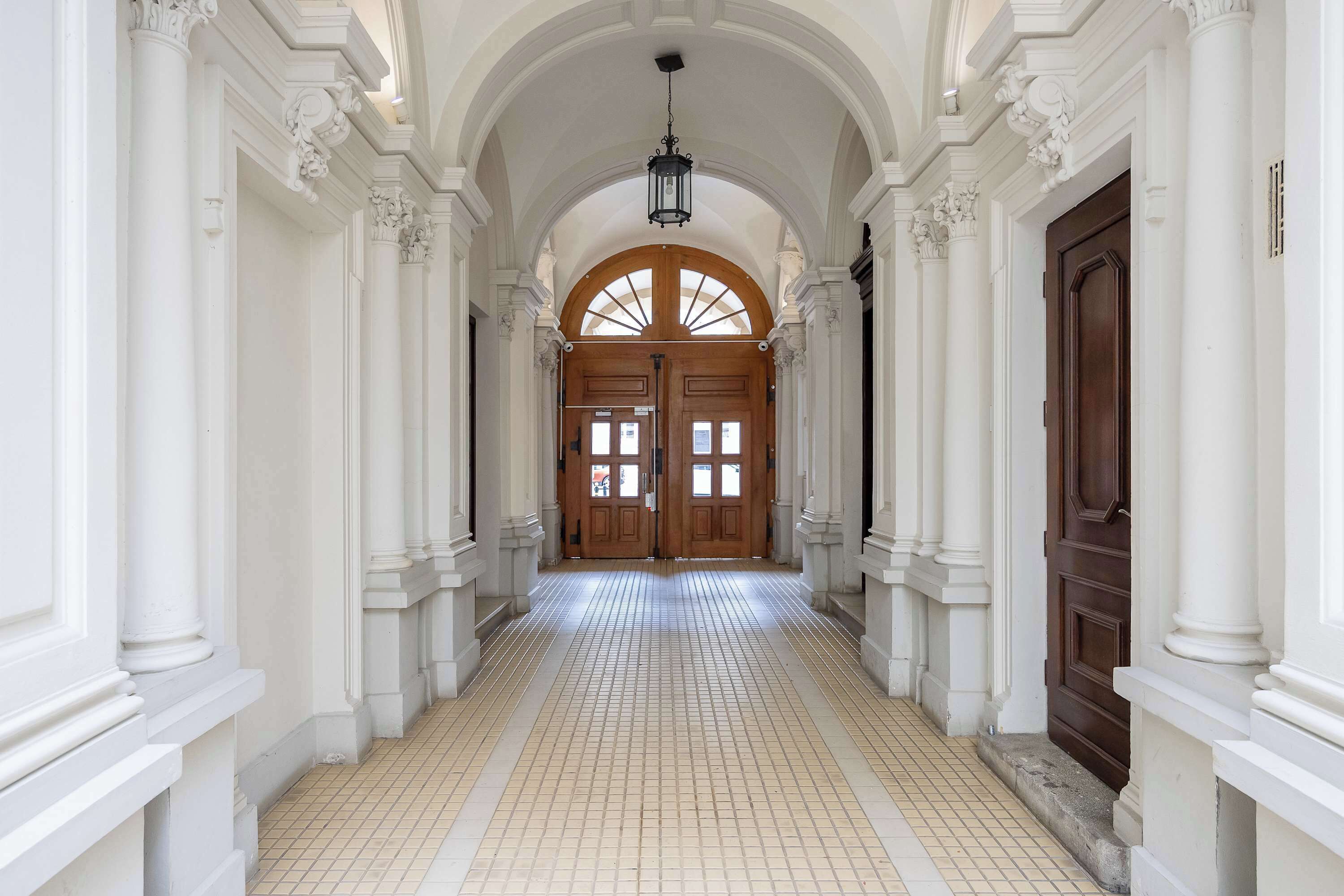 Un couloir aux colonnes blanches, au sol carrelé, et une porte en bois au fond.