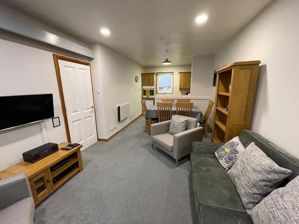 A compact living space with a flat-screen TV, a sofa, a dining table with chairs, and a kitchen area.