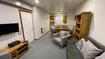 A compact living space with a flat-screen TV, a sofa, a dining table with chairs, and a kitchen area.
