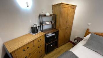 A bedroom with a wooden dresser, a wardrobe, a bed with a grey blanket, and a bedside table with a lamp.