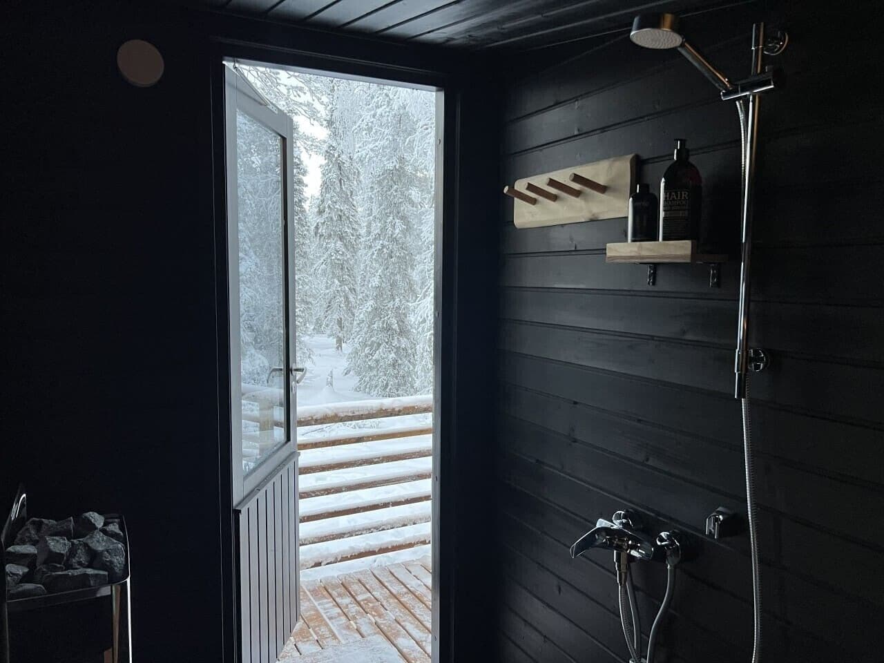 Un baño con puerta de madera que da a un área exterior nevada, ducha y estante con artículos de aseo.