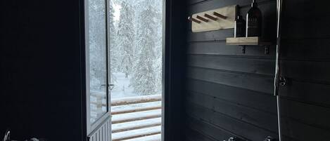 Un baño con puerta de madera que da a un área exterior nevada, ducha y estante con artículos de aseo.
