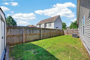 A backyard with a wooden fence, a grassy lawn, and a house with a sloped roof. - Roadmap Lodging: 3BR + Garage Near Regional Health (Columbus)