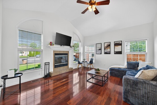 A spacious living room with hardwood flooring, a fireplace, a flat-screen TV, and a ceiling fan.