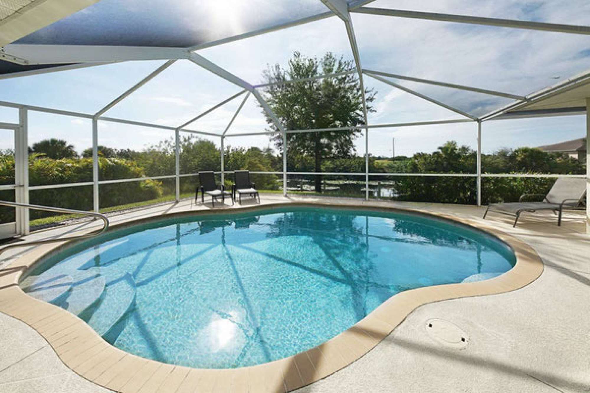 A clear swimming pool with a covered area, surrounded by lounge chairs and a view of trees.