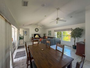 Uma sala de estar ampla com uma mesa de jantar, sofás, ventiladores de teto e uma grande janela com vista para áreas verdes.
