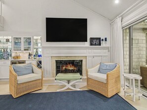 Ein Wohnzimmer mit Kamin, zwei Korbsesselsofas, einem Flachbildfernseher und einer Küchenzeile mit Glastür.