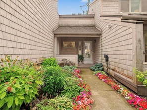 A narrow pathway flanked by lush greenery and vibrant flowers leading to a house entrance with a visible door and windows. - Beautiful Breakers East Condo with Lake Michigan Views (Harbor Springs)