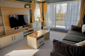 A modern living room with a flat-screen TV, a coffee table, a sofa, and a large window with curtains. - Kielder - Spacious 3 Bed, Luxury Lodge, Haven, Seton Sands (Port Seton)