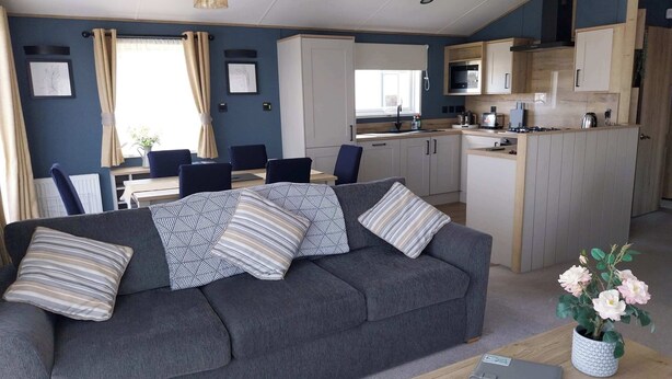 A modern living room with a grey sofa, a dining area with a table and chairs, and a kitchen with wooden cabinets and appliances. - Kielder - Spacious 3 Bed, Luxury Lodge, Haven, Seton Sands (Port Seton)