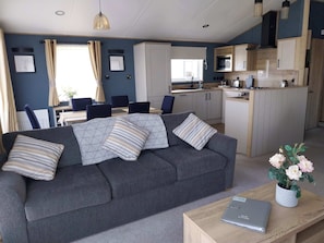 A modern living room with a grey sofa, a dining area with a table and chairs, and a kitchen with wooden cabinets and appliances. - Kielder - Spacious 3 Bed, Luxury Lodge, Haven, Seton Sands (Port Seton)