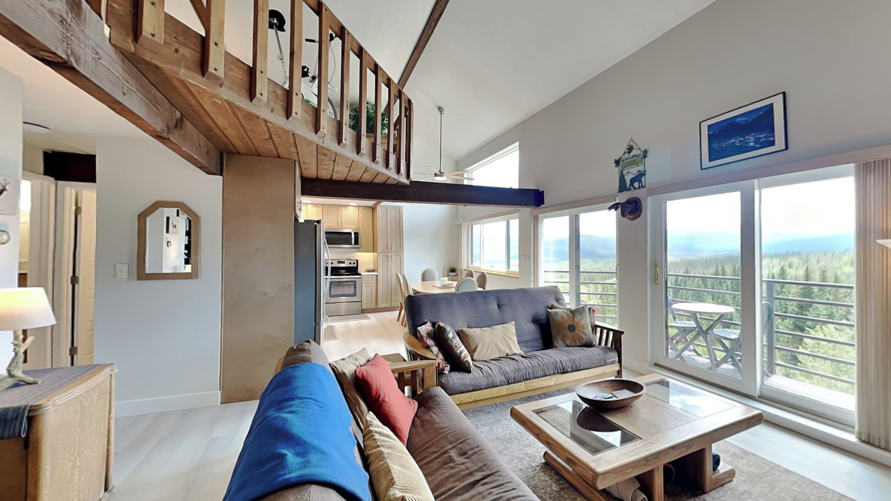A spacious living area with a high ceiling, a wooden staircase, a sofa set, a dining table, and a large window offering a scenic view.