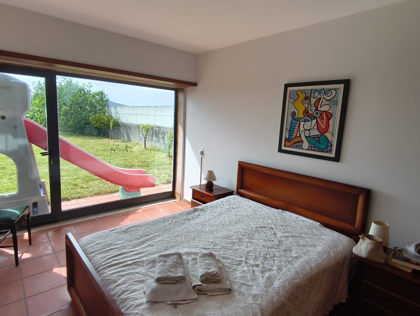A bedroom with a bed, a nightstand, a lamp, a painting, and a view of a slide and garden through a large window.