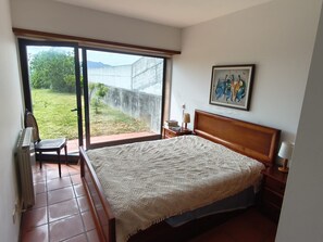 A bedroom with a bed, a nightstand, a lamp, a framed picture, and a view of a garden through a sliding door.