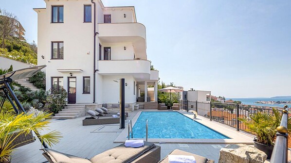 A modern two-story building with a swimming pool, outdoor seating, and a view of the sea.