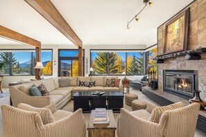 A spacious living room with a large sectional sofa, a fireplace, and a view of autumn foliage through large windows. - Panoramic rooftop deck, captivating views, and tranquil luxury (Dillon)