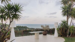 Un patio al aire libre con una mesa y sillas, rodeado de plantas tropicales y vistas a la costa.