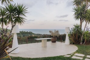 An outdoor patio with a table and chairs, surrounded by tropical plants and overlooking a coastal view. - LA GRIGLIA DI CASA NOSTRA SRL (Silvi)