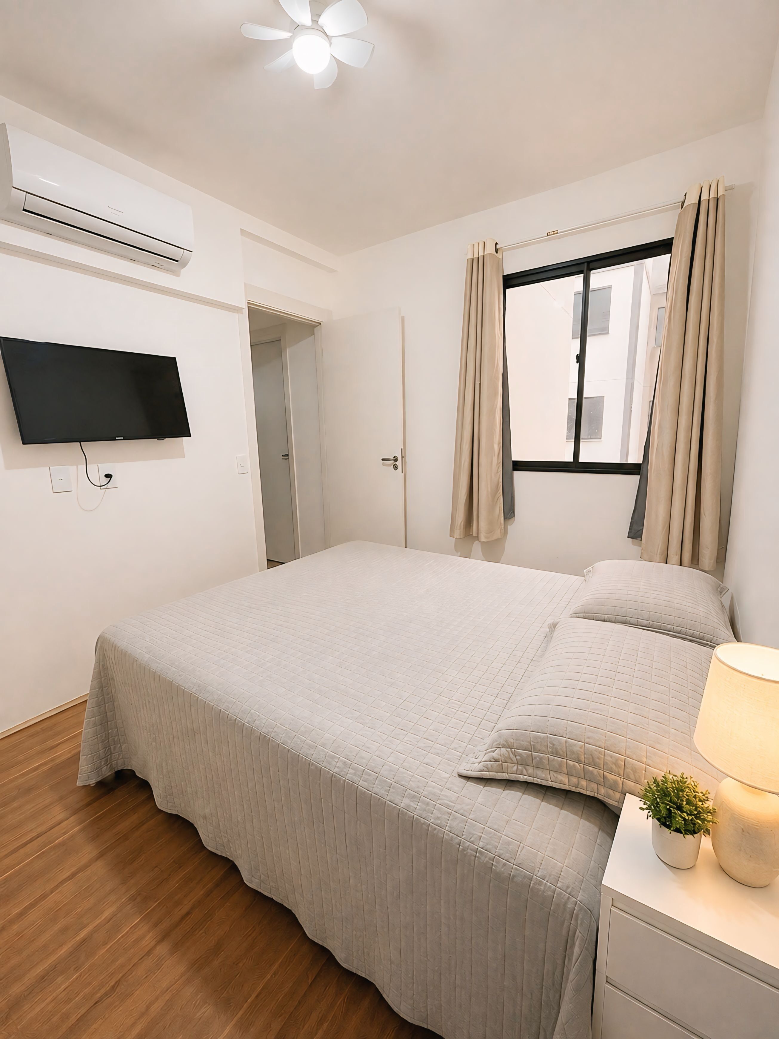 Une chambre à coucher avec un lit, un téléviseur fixé au mur, une fenêtre avec des rideaux et une table de chevet avec une lampe et une plante.