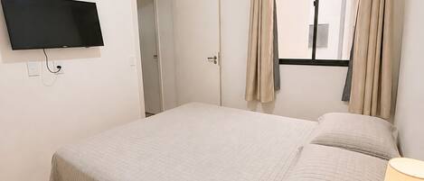 A bedroom with a bed, a TV mounted on the wall, a window with curtains, and a bedside table with a lamp and a plant.