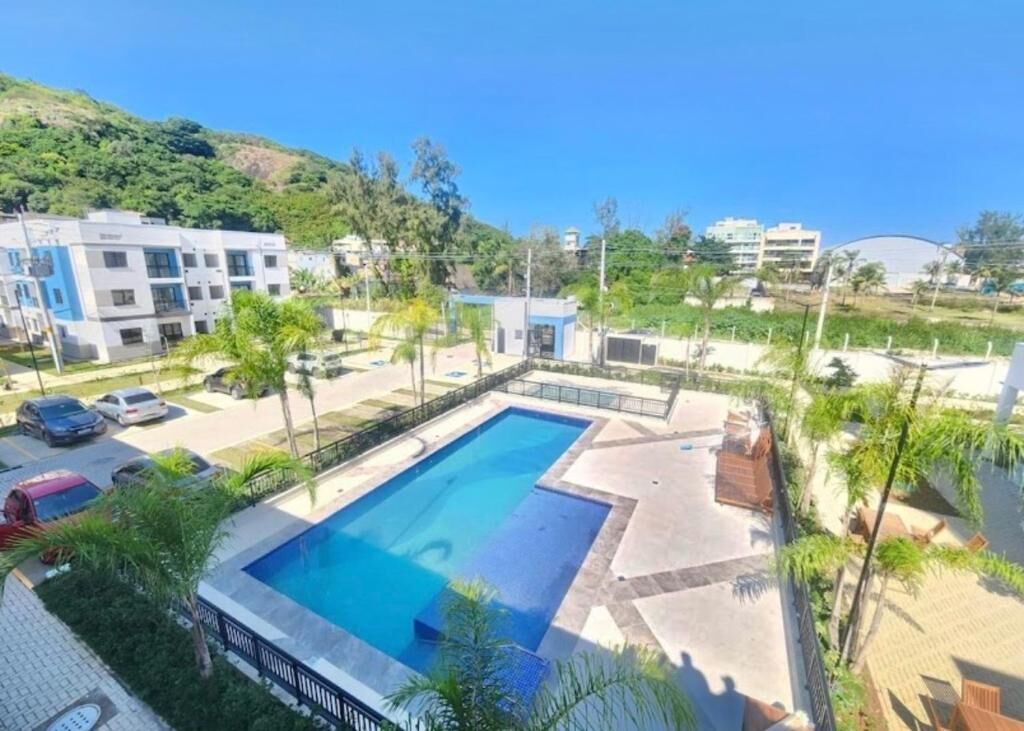 A swimming pool surrounded by a paved area with palm trees, a parking lot, and buildings in the background.