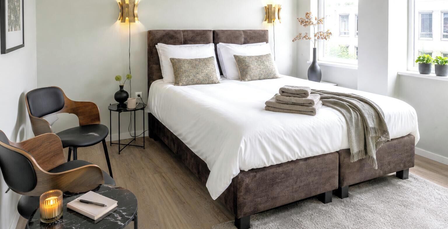 A bedroom with a large bed, bedside tables, a chair, a small table with a candle, and a window with plants.