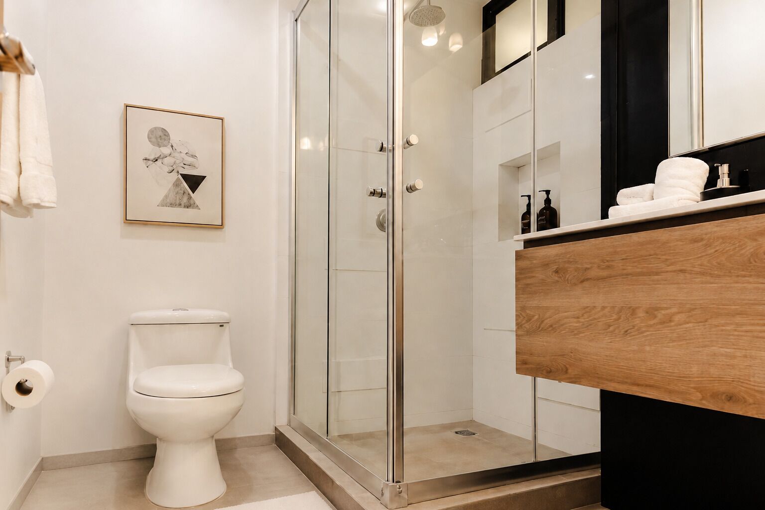 Une salle de bain moderne avec une douche à parois de verre, des toilettes blanches, un meuble-lavabo en bois et un tableau encadré au mur.