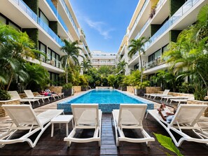Zona de piscina con sillones de descanso y una piscina azul cristalina, rodeada de follaje y arquitectura moderna.