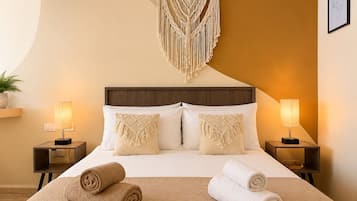A hotel room with a bed, two bedside lamps, a macrame wall hanging, and two rolled towels on the bed.