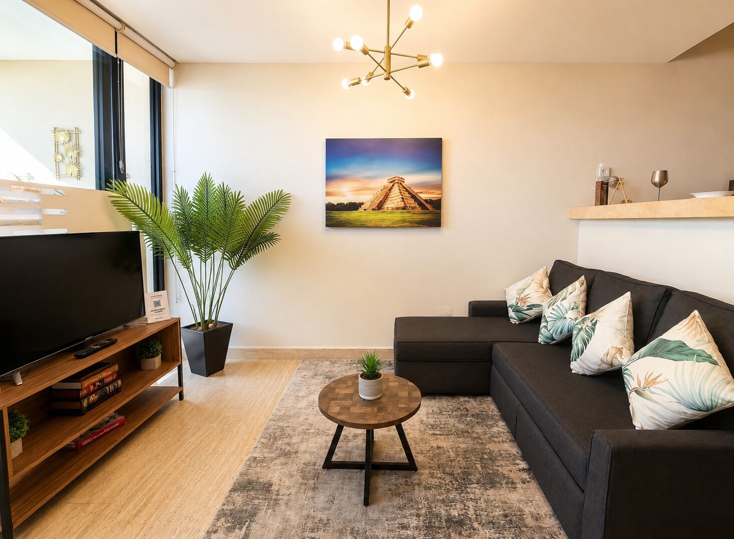 A modern living room with a flat-screen TV, a dark sofa with decorative pillows, a round coffee table, a potted plant, and a painting of a pyramid on the wall.