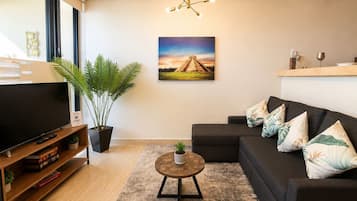A modern living room with a flat-screen TV, a dark sofa with decorative pillows, a round coffee table, a potted plant, and a painting of a pyramid on the wall.