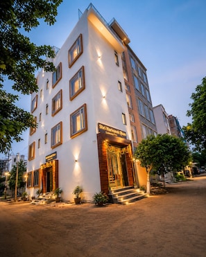 A multi-story hotel with a white facade, multiple windows, and a well-lit entrance. - SHRI HARIVANSH DHAM (Mathura)