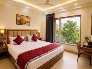 A hotel room with a large bed, a desk, a chair, a ceiling fan, a window with a view, and a painting on the wall. - SHRI HARIVANSH DHAM (Mathura)