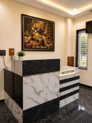 A reception desk with a marble finish, a framed religious painting above it, and a window with blinds to the right. - SHRI HARIVANSH DHAM (Mathura)